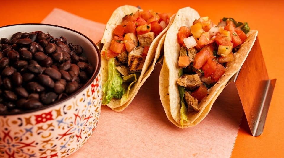 Two tacos with meat, lettuce, and salsa, next to a bowl of black beans.