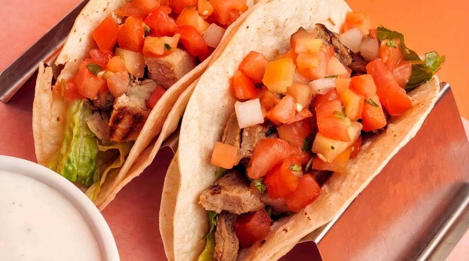 Two tacos with grilled meat, lettuce, and tomato salsa, with a side of white sauce.