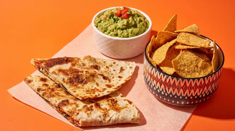 Two quesadilla halves, a bowl of guacamole, and a bowl of tortilla chips on an orange background.