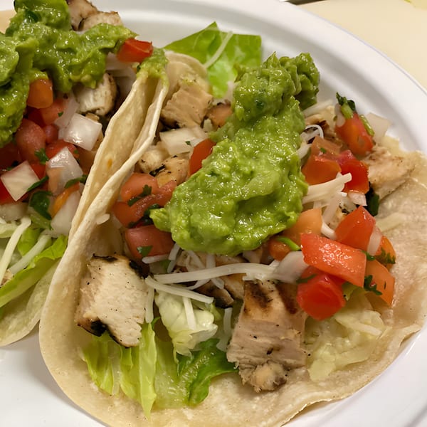 Two chicken tacos with lettuce, pico de gallo, and guacamole on a white plate.