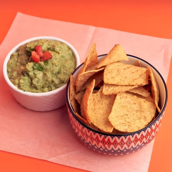 A bowl of guacamole topped with diced tomatoes and a bowl of tortilla chips on an orange background.