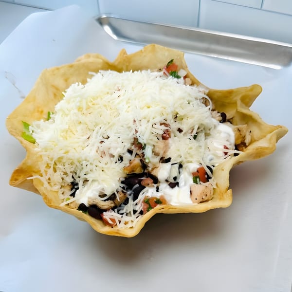 A taco salad in a crispy tortilla bowl, topped with shredded cheese, chicken, and black beans.