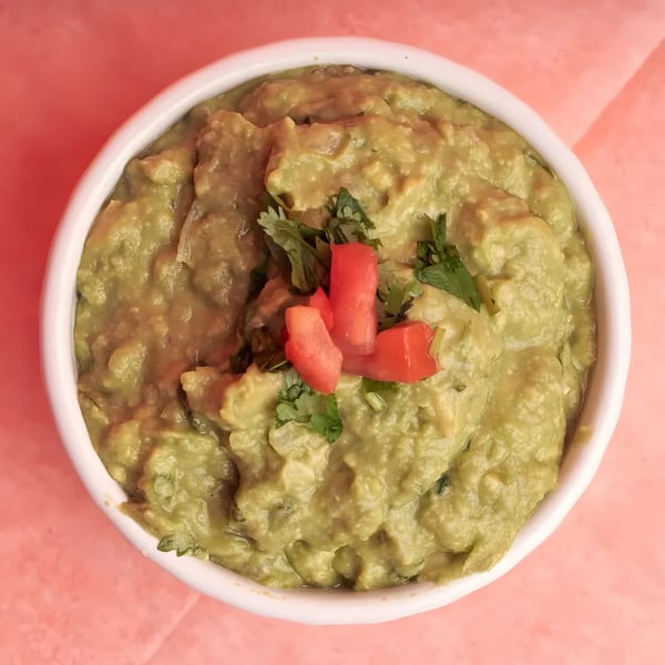 A white bowl filled with chunky guacamole, garnished with cilantro and diced tomatoes, on a pink surface.